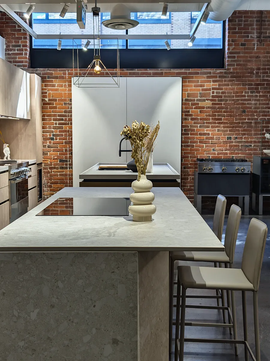 A pale stone kitchen island with a sculptural vase, bar stools and an exposed brick wall behind.