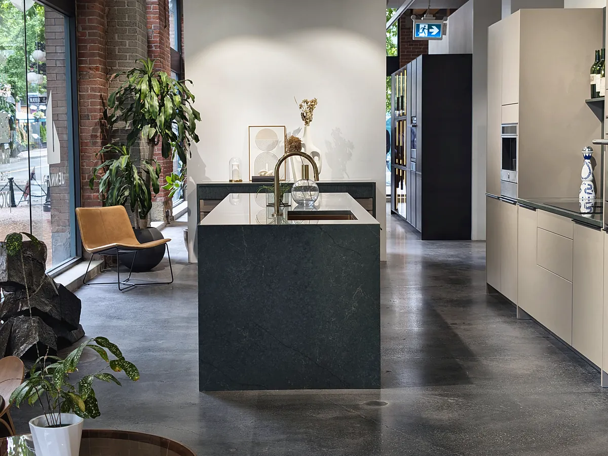 A green stone kitchen island with pale cabinetry beside tall street windows, a leather chair and plants.